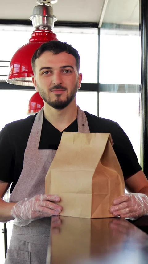 Vertical video of friendly young guy in apron holding paper package with take Stock Footage 280386478