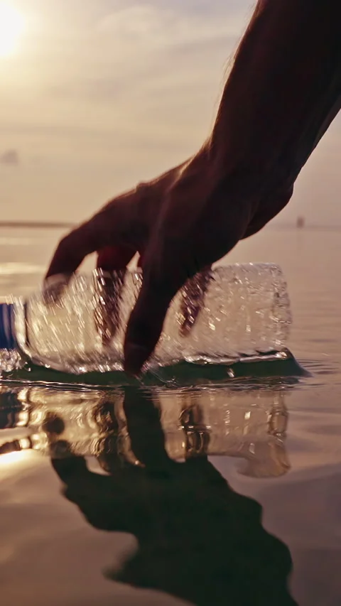 Vertical video. Hand Lifting Crushed Plastic Bottle from Calm Sea at Sunset with Stock Footage 329059708