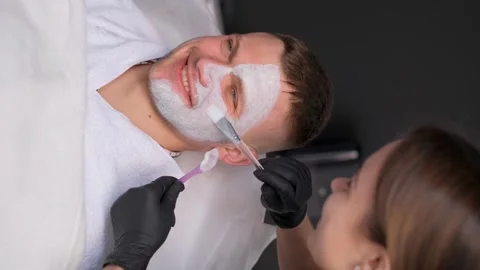 Vertical video of a handsome, smiling man applying a face mask to a beautician Stock Footage 250356363