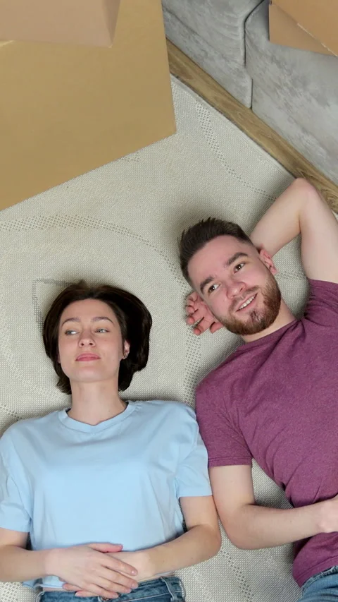 Vertical video. Happy couple lying on the floor in new home with cordboard boxes Stock Footage 346201874