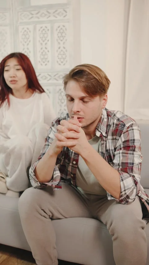 Vertical video A husband sits on the couch, looking sad and puzzled, as his Stock Footage 295549355