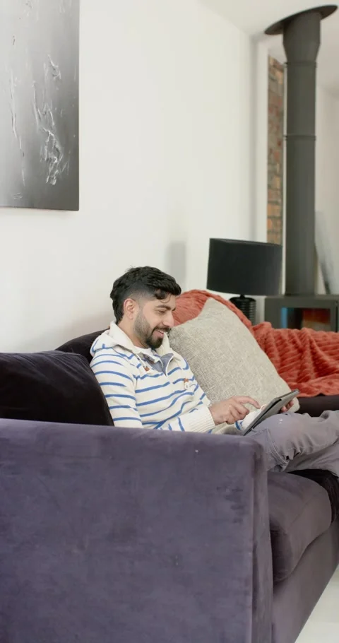 Vertical video: Indian man leaning on sofa tapping, scrolling tablet in living Stock Footage 345395783