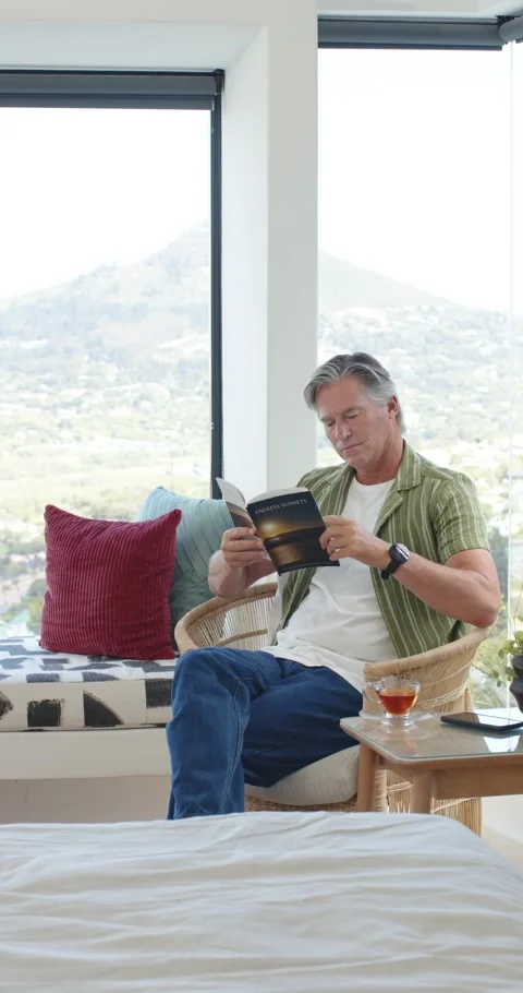 Vertical video: Man in 50s reading booklet in armchair by window, phone and tea Stock Footage 346724106