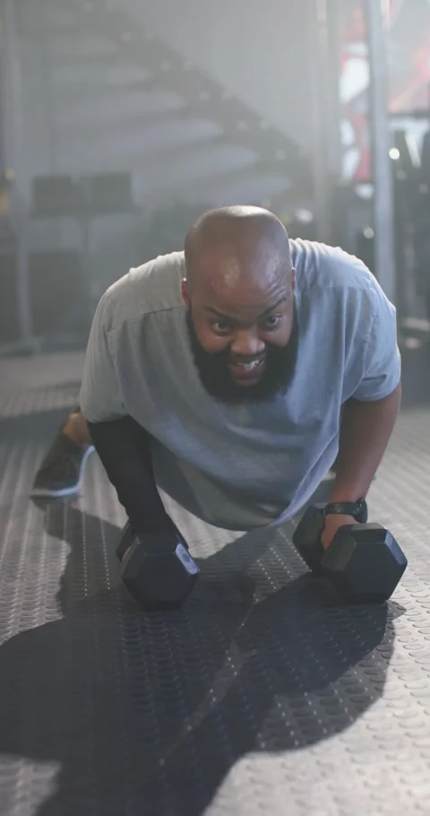 Vertical video: Man bracing, doing push-ups and single-arm rows on hex dumbbells Stock Footage 345130261