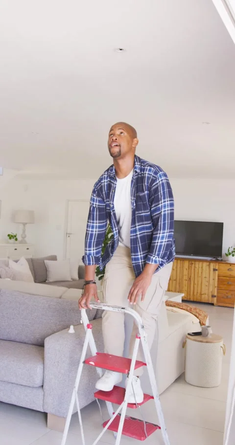 Vertical video: Man bringing step ladder near sofa, climbing and fixing ceiling Video stock 345492928