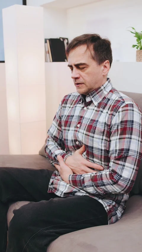 Vertical video Man experiencing abdominal pain while sitting on a couch in a Stock Footage 301581790