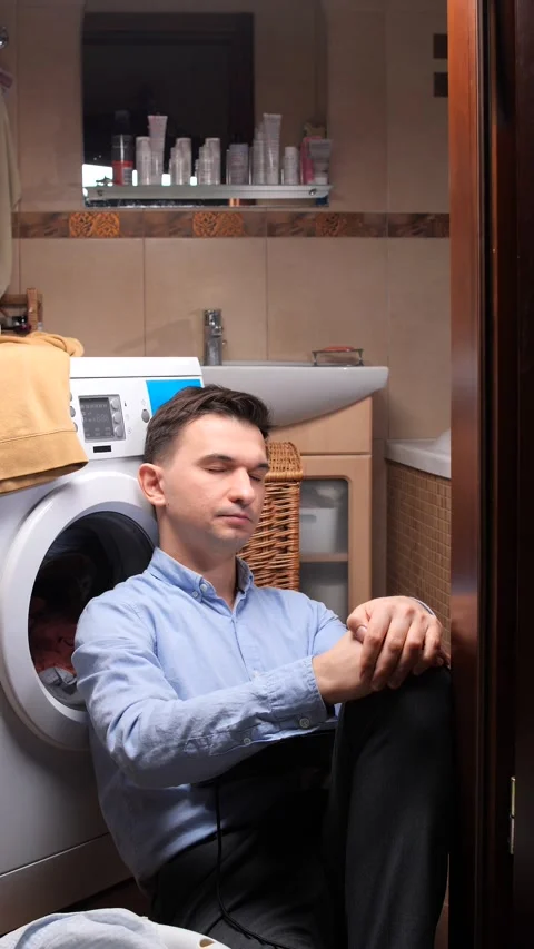 Vertical video man leaning against a washing machine, trying to work on his Stock Footage 323974886