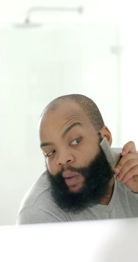 Vertical video: Man leaning to mirror, lifting trimmer and grooming beard while Stock Footage 346724246