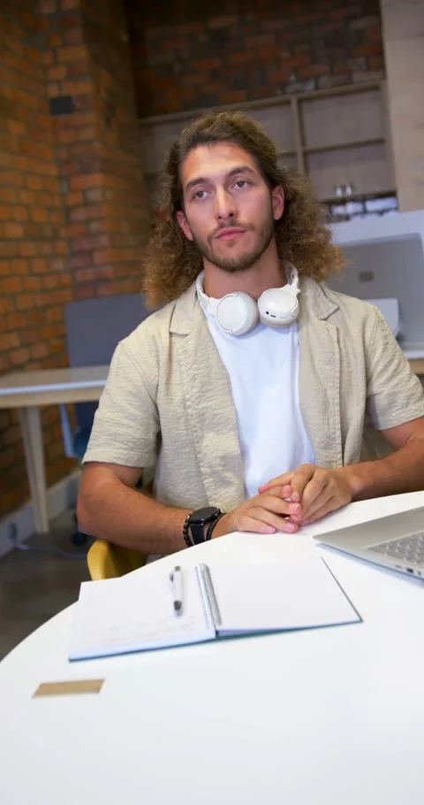 Vertical video: Man mid20s sitting at laptop rubbing jaw, putting on headphones, Stock Footage 342035839