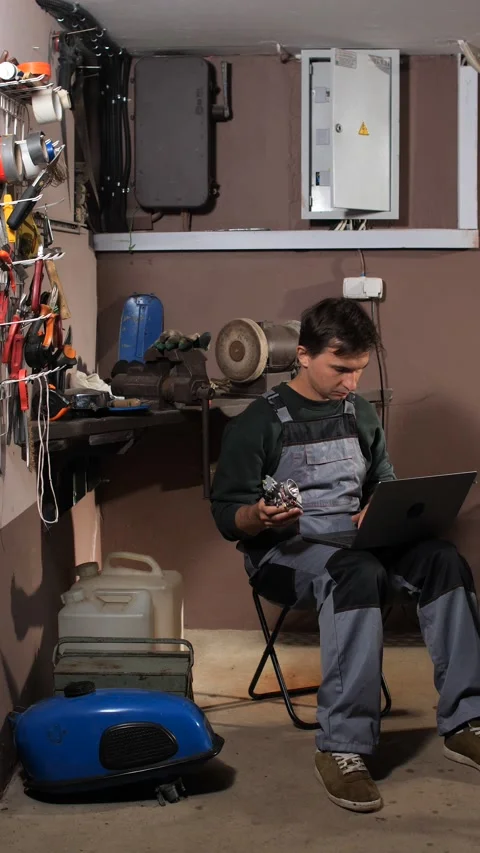 Vertical video man in overalls sitting in a workshop, holding a small engine Stock Footage 342773798