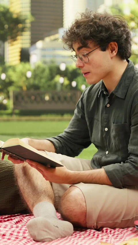 Vertical Video Man reading poetry book aloud to woman laying on checkered picnic Stock Footage 341472670