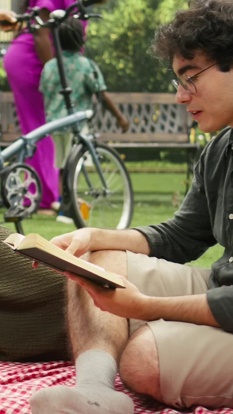 Vertical Video Man reading poetry book aloud to woman laying on checkered picnic Stock Footage 341473360