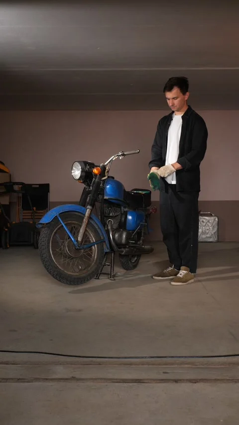 Vertical video man standing by his classic 1970s blue motorcycle, holding work Stock Footage 342760711