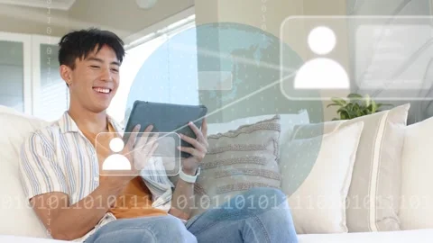 Vertical video: Man tapping tablet on sofa, forming globe, icons and binary Stock Footage 332772341