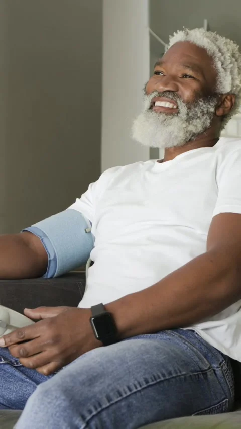 Vertical video: Pressing button, senior in white tee checking BP on couch for Stock Footage 348203190