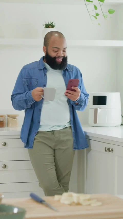 Vertical video: Reading alert, man in denim at kitchen counter, sipping mug, Stock Footage 348728344