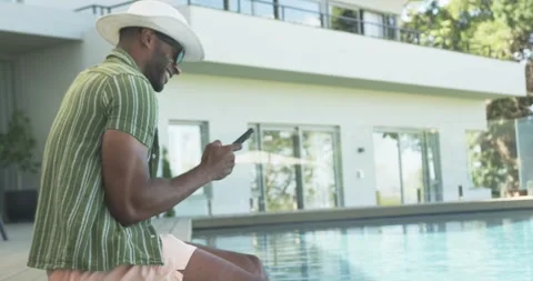 Vertical video: Reading phone man in trunks donning hat dipping feet in pool Stock Footage 340987632