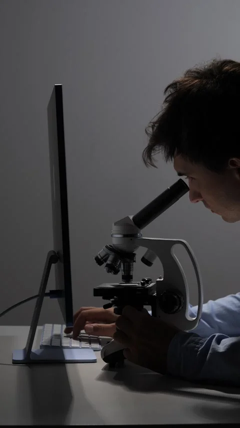 Vertical video scientist looking into a microscope while typing on a computer Stock Footage 342673565