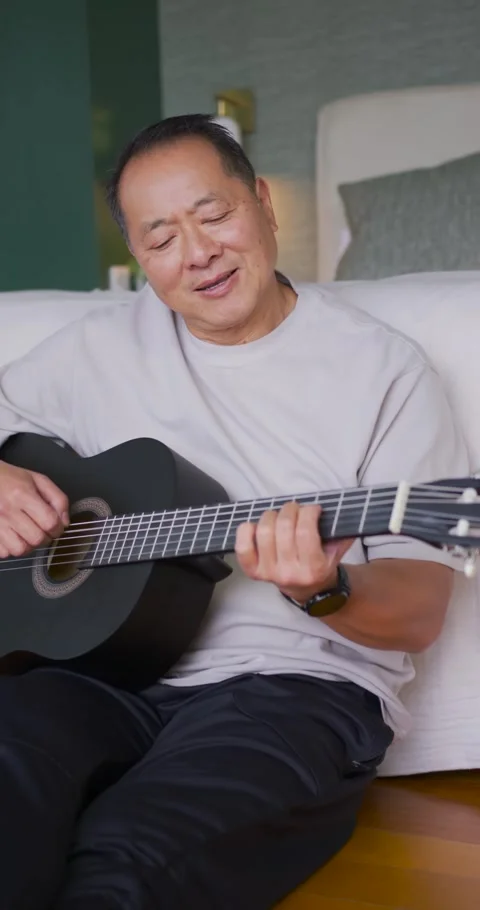 Vertical video: Senior Asian man sitting at home on floor with black guitar Stock Footage 337734336