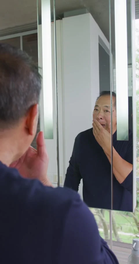 Vertical video: Senior Asian man inspecting mirror at vanity, rubbing cheeks and Stock Footage 337734353