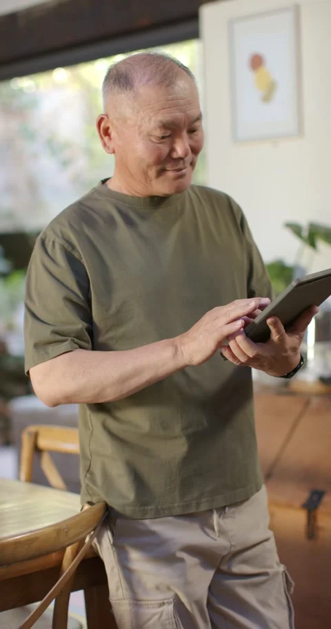 Vertical video: Senior Asian man standing near dining table holding tablet, Stock Footage 345323638