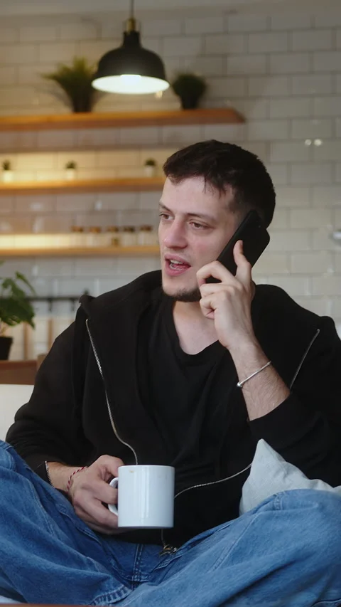Vertical video. Smiling young man sitting on the sofa talking on his mobile.. Stock Footage 273394243