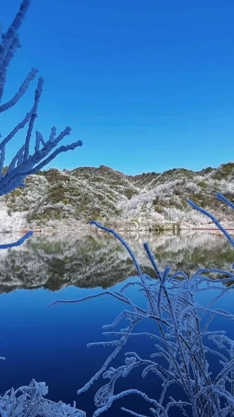 Vertical video of snowy hills reflected in calm deep blue lake with rime ice Video stock 327582483