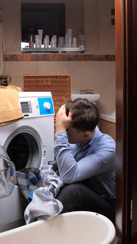 Vertical video stressed man crouching by an open washing machine, holding his Stock Footage 324761338