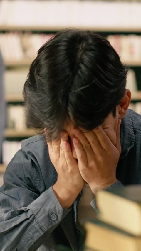 Vertical Video Tired asian student leaning over open book between tall stacks in Stock Footage 336380749