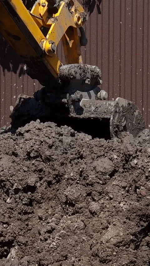 Vertical video. A tractor bucket digs a hole near the brown fence of a house. Stock-Footage 281540106