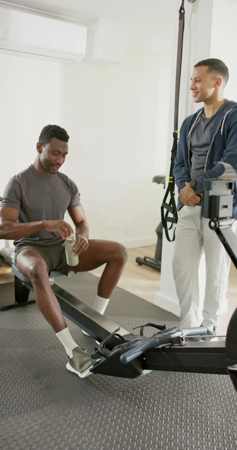 Vertical video: Trainer guiding Black man on rowing machine sipping water and Stock-Footage 345129410