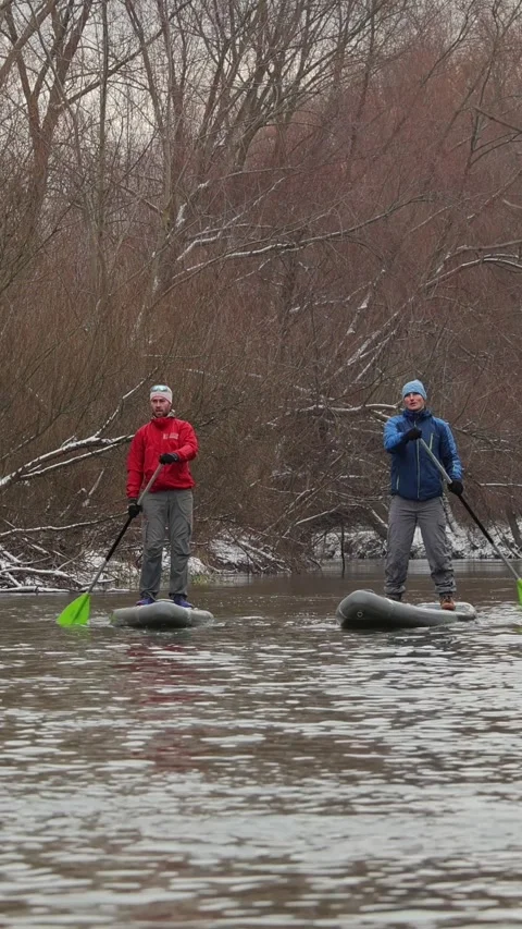 Vertical video. Two mans paddleboarding in a tranquil river with snowy trees Video stock 299617838