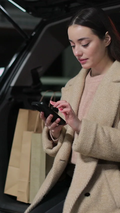 Vertical video Woman using a smartphone while sitting in the trunk of a car Stock Footage 339574708