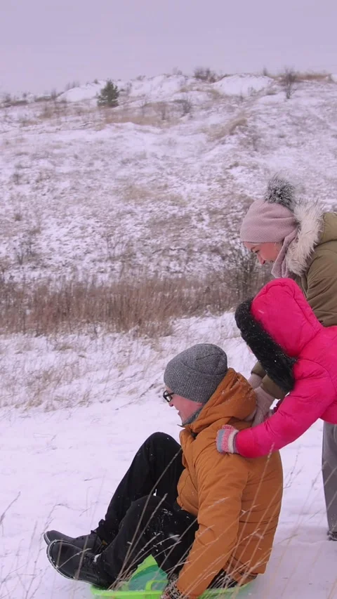 Vertical video. A young father gets on a green sled and is pushed down a hill by Video stock 301554042
