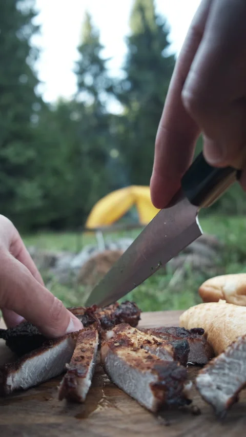 Vertical video of young man cutting hot beef steak just cooked on bonfire Stock Footage 288014329
