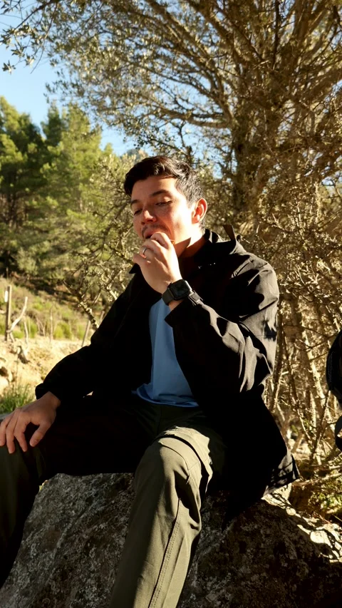 Vertical video of young man sitting on a mountain eating a sandwich looking.. Stock Footage 296164589