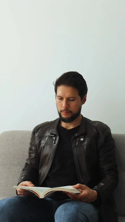 Vertical view of a concentrating man reading a text book sitting on a grey couch Stock Footage 340583297