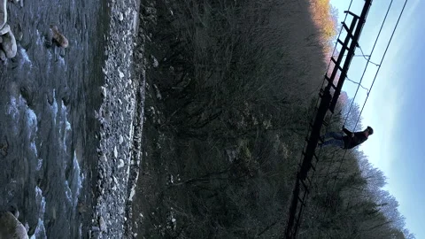 Vertical: young man carefully walks on a suspension wooden bridge over a river Video stock 233345488