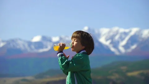A very cute boy drinks orange juice on a background of beautiful mountains. 4k Stock Footage 83236432