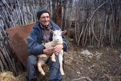 A very old man in messy clothes is sitting on a stool in the yard of an old f Stock Photos
