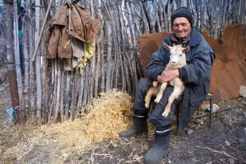 A very old man in messy clothes is sitting on a stool in the yard of an old f Stock Photos