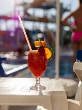 A vibrant cocktail with fruit garnishes sits on a white table near a pool. Br Stock Photos