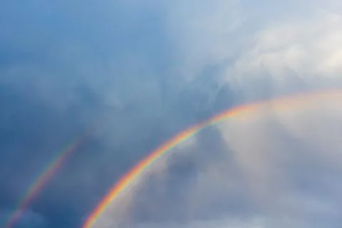 A vibrant double rainbow arcs across a moody blue sky with soft clouds Stock Photos