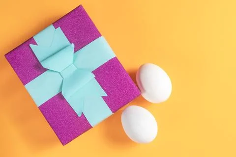 Vibrant gift box with a blue bow and two white eggs on an orange background Stock Photos