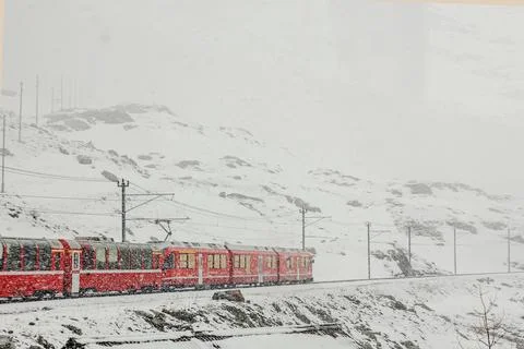 A vibrant red train makes its way through a snowy mountain landscape. Stock Photos