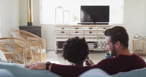 Video of back view of happy diverse couple drinking coffee and watching tv with Stock Footage 341851299