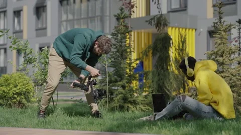 Video maker with professional video camera shoots african sitting on lawn in Stock Footage 244268551