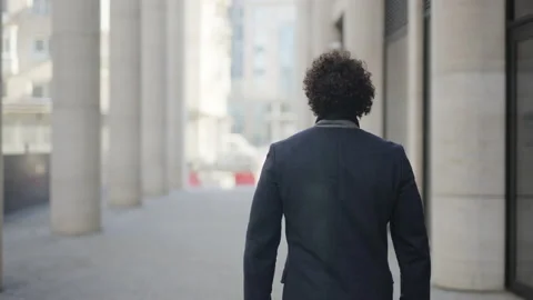 View from the back: a man with dark curly hair walks forward, looks at his watch Stockbeeldmateriaal 200139562