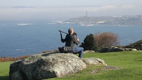 View from behind of bagpiper sitting on a rock playing the bagpipes Stock Footage 148001033
