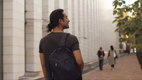 View from behind of a young man with a backpack walks alongside the building Stock Footage 253971884